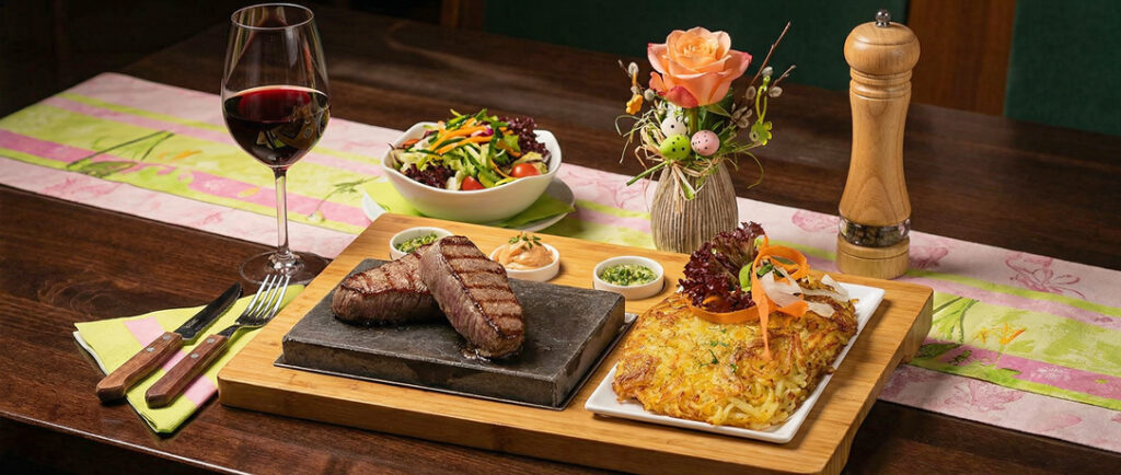 Heißer Stein mit zwei gegrillten Rindersteaks, Rösti, frischem Salat, Dips und einem Glas Rotwein auf einem festlich gedeckten Holztisch.