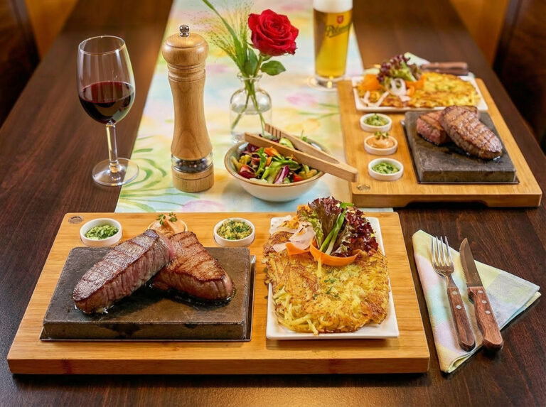 Heißer Stein mit zwei gegrillten Rindersteaks, Rösti, frischem Salat, Dips und einem Glas Rotwein und Bier auf einem festlich gedeckten Holztisch.