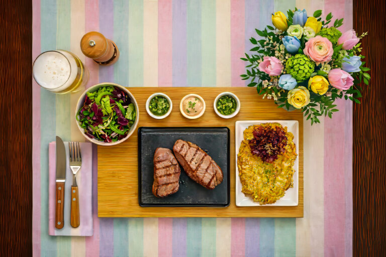 Heißer Stein mit zwei gegrillten Rindersteaks, Rösti, frischem Salat, Dips und einem Glas Rotwein auf einem festlich gedeckten Holztisch.